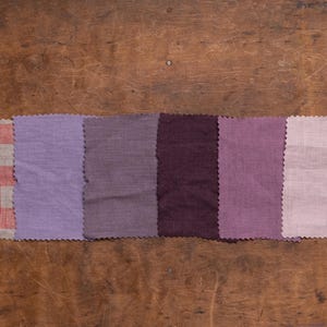 Linen fabric pieces in pink, purple, and plum tones shown in a flat lay detail view, lined up in a single horizontal row on worn wood, with pinked edges, one checkered swatch, and five plain woven samples, Knock Knock Linen.