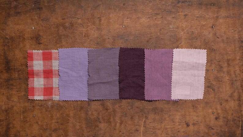 Linen fabric pieces in pink, purple, and plum tones shown in a flat lay detail view, lined up in a single horizontal row on worn wood, with pinked edges, one checkered swatch, and five plain woven samples, Knock Knock Linen.