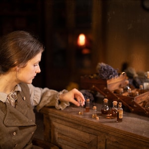 Brown linen vest on seated model, side-profile waist-up; worn over light gray blouse with ruffled collar and tie; front has wide flap with buttonholes and small buttons; her hand reaches toward corked glass vials on a wooden cabinet.