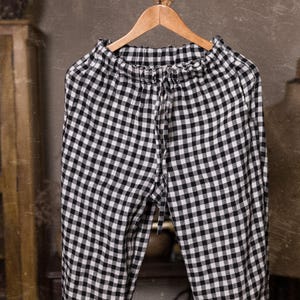 Black and white checked linen pants, shown hanging in a close-up front-facing photo, with a ruffled elastic waist, long front drawstring, softly wrinkled fabric, and a wooden hanger beneath a rustic branch, Knock Knock Linen.