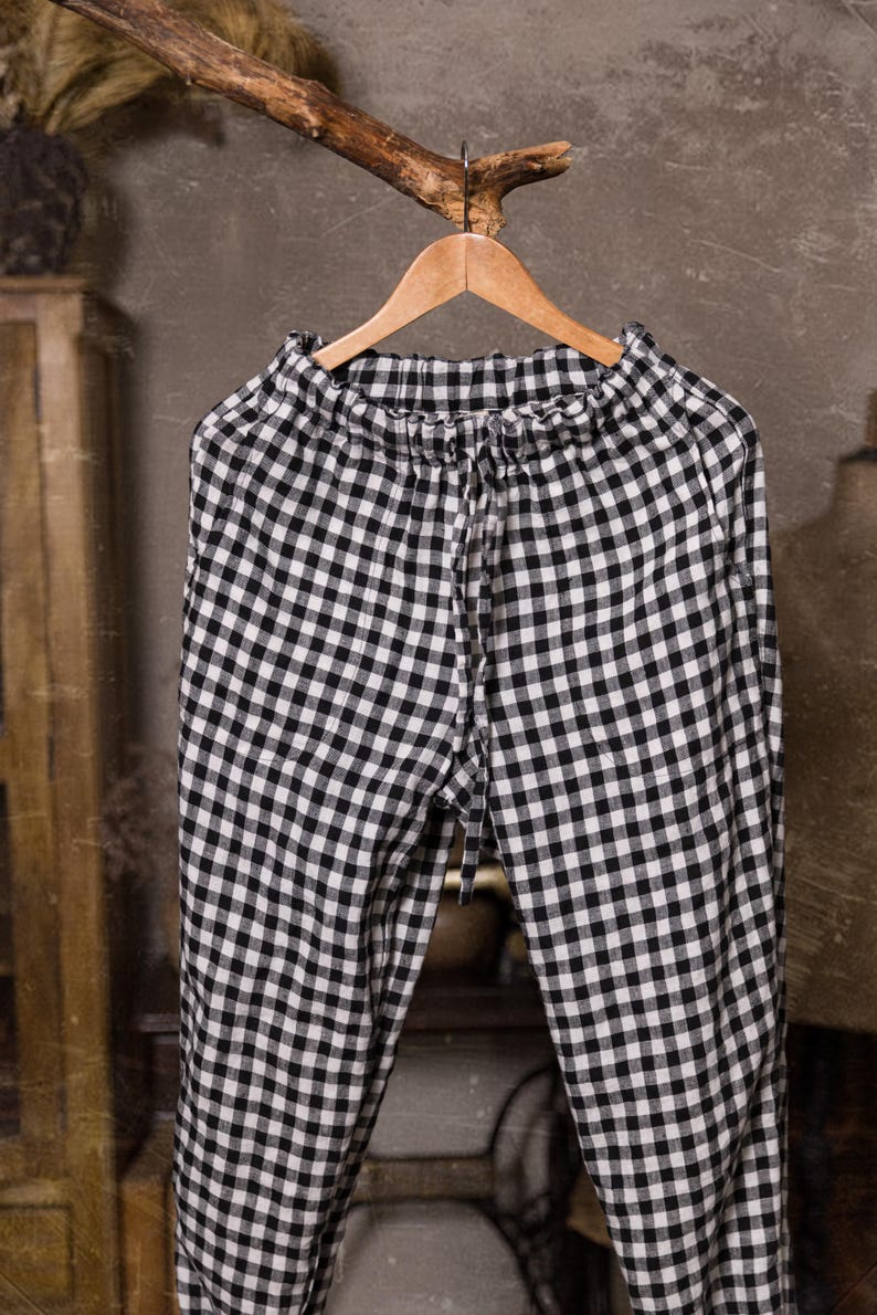 Black and white checked linen pants, shown hanging in a close-up front-facing photo, with a ruffled elastic waist, long front drawstring, softly wrinkled fabric, and a wooden hanger beneath a rustic branch, Knock Knock Linen.