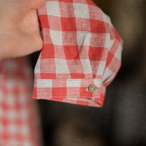 Close-up of a hand holding the cuff of a red and white gingham cottage rose linen dress. Cuff features a single beige button. Lightweight woven fabric with checkered pattern. Background shows matching fabric, suggesting full outfit.