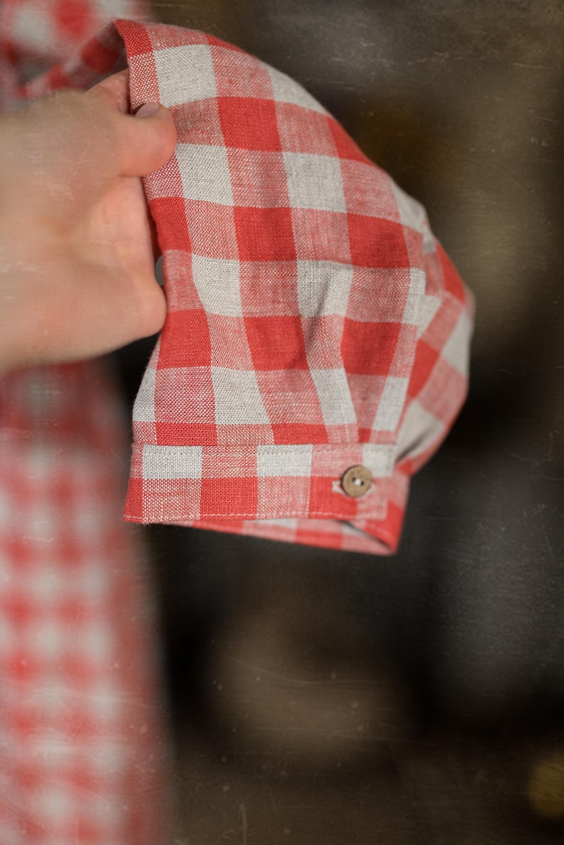 Close-up of a hand holding the cuff of a red and white gingham cottage rose linen dress. Cuff features a single beige button. Lightweight woven fabric with checkered pattern. Background shows matching fabric, suggesting full outfit.