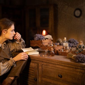 SPARROW linen vest in brown, displayed on a seated model in a side-profile mid-length view, with a sleeveless shape, rounded front edge, visible front buttons, over a light gray blouse with a tie neckline, beside an open notebook and glass bottles, Knock Knock Linen.