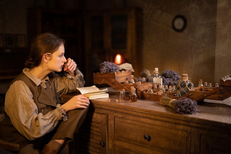 SPARROW linen vest in brown, displayed on a seated model in a side-profile mid-length view, with a sleeveless shape, rounded front edge, visible front buttons, over a light gray blouse with a tie neckline, beside an open notebook and glass bottles, Knock Knock Linen.