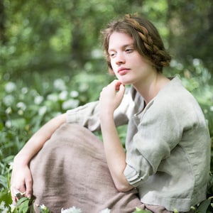 Woman seated outdoors among green foliage and wildflowers, wearing a loose-fitting natural herringbone linen blouse with a boat neck and gathered cuffs, paired with a brown skirt. Shown on model in a relaxed pose. Linen blouse, romantic top women.