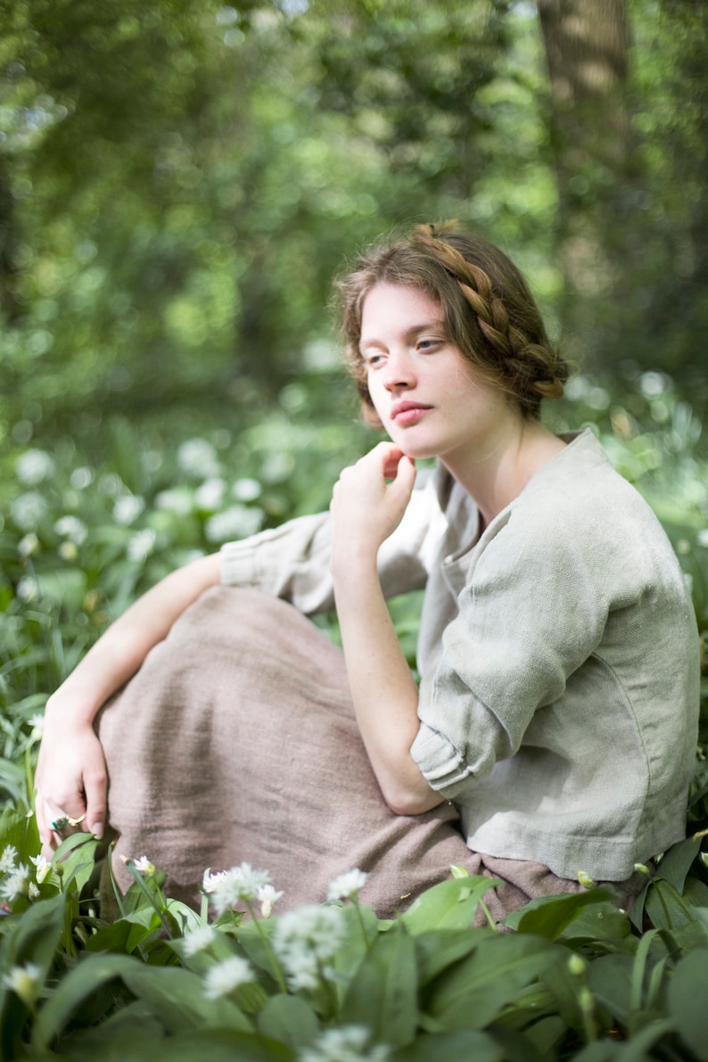 Woman seated outdoors among green foliage and wildflowers, wearing a loose-fitting natural herringbone linen blouse with a boat neck and gathered cuffs, paired with a brown skirt. Shown on model in a relaxed pose. Linen blouse, romantic top women.