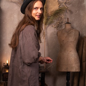 Woman wearing one loose-fit brownish-gray linen boho dress with short sleeves and pockets, styled with black wide-brim hat. Standing beside vintage dress form. Indoor setting with dried palm leaves and candle. Linen summer dress, boho witch clothing.