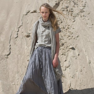 Model standing barefoot on sand wearing frost grey linen maxi skirt with drawstring waist, side pockets, and elastic waistband; paired with matching short-sleeved top and scarf. Fabric has crinkled texture. Keywords: linen skirt, maxi skirt, elastic waist skirt.