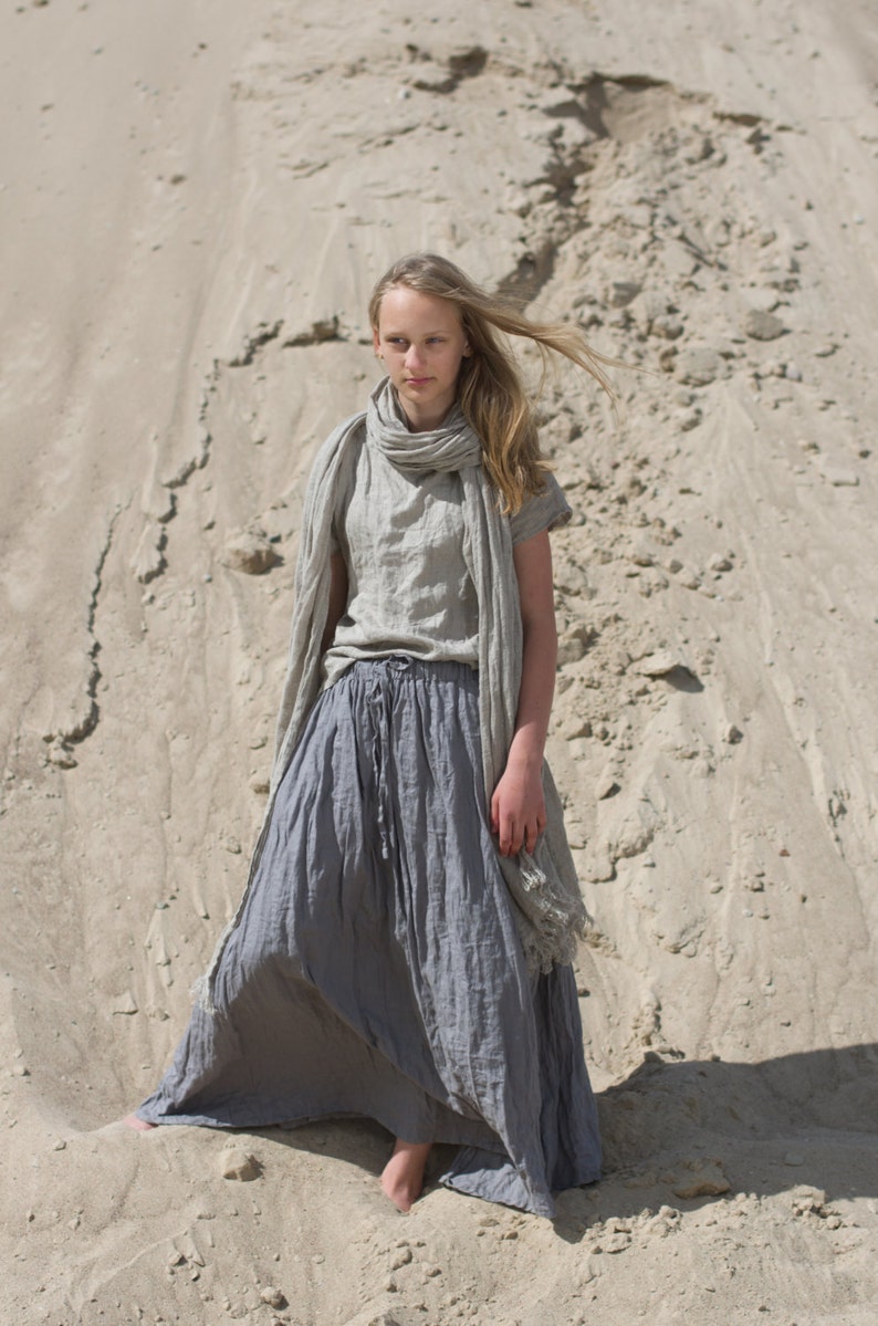 Model standing barefoot on sand wearing frost grey linen maxi skirt with drawstring waist, side pockets, and elastic waistband; paired with matching short-sleeved top and scarf. Fabric has crinkled texture. Keywords: linen skirt, maxi skirt, elastic waist skirt.
