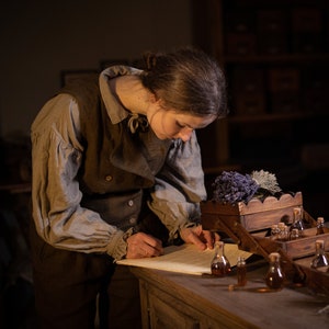 SPARROW linen vest in brown, shown on a model in a side three-quarter mid-length photo, with a sleeveless cut, curved overlapping front flap, light buttons, and a fitted shape, over a light gray collared blouse at a wooden table, Knock Knock Linen.