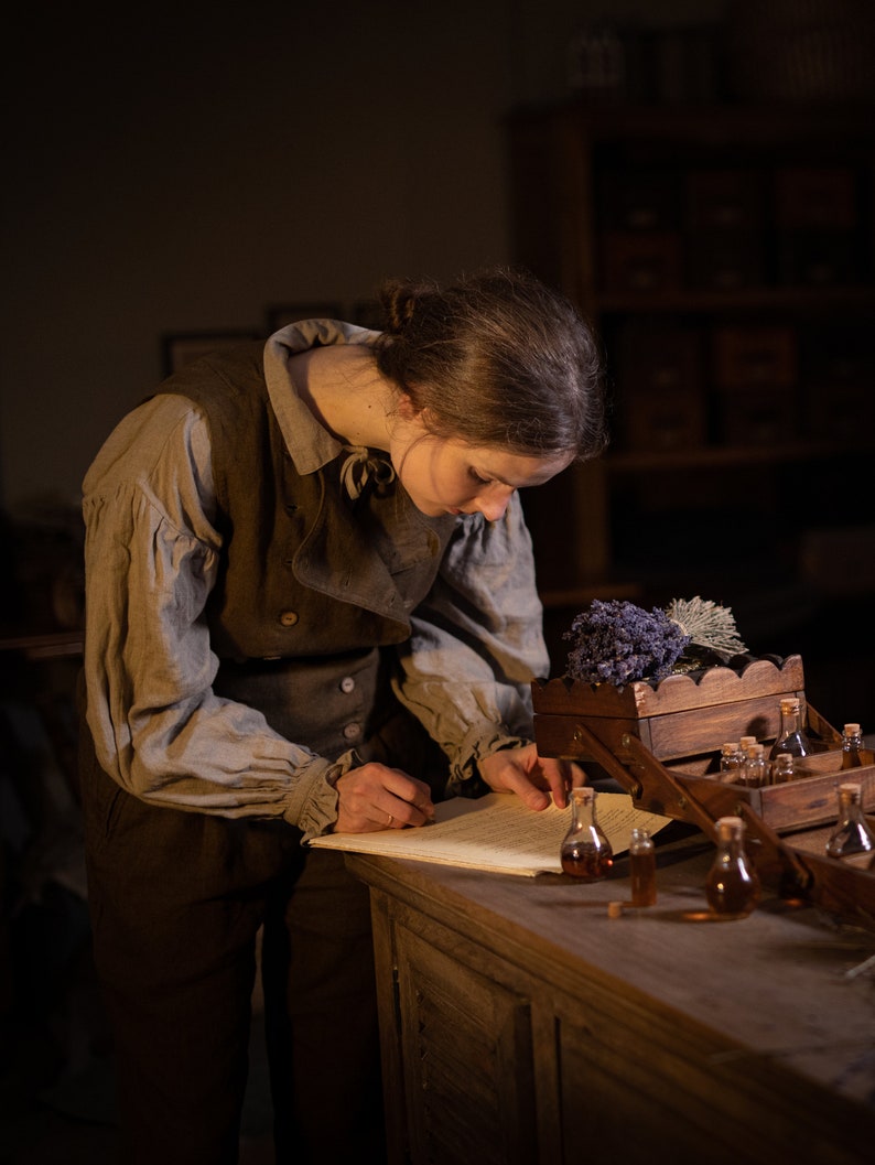 SPARROW linen vest in brown, shown on a model in a side three-quarter mid-length photo, with a sleeveless cut, curved overlapping front flap, light buttons, and a fitted shape, over a light gray collared blouse at a wooden table, Knock Knock Linen.