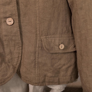 A close-up of a brown linen jacket with two wooden buttons and a small pocket. The jacket has a tailored fit and a natural, textured appearance.
