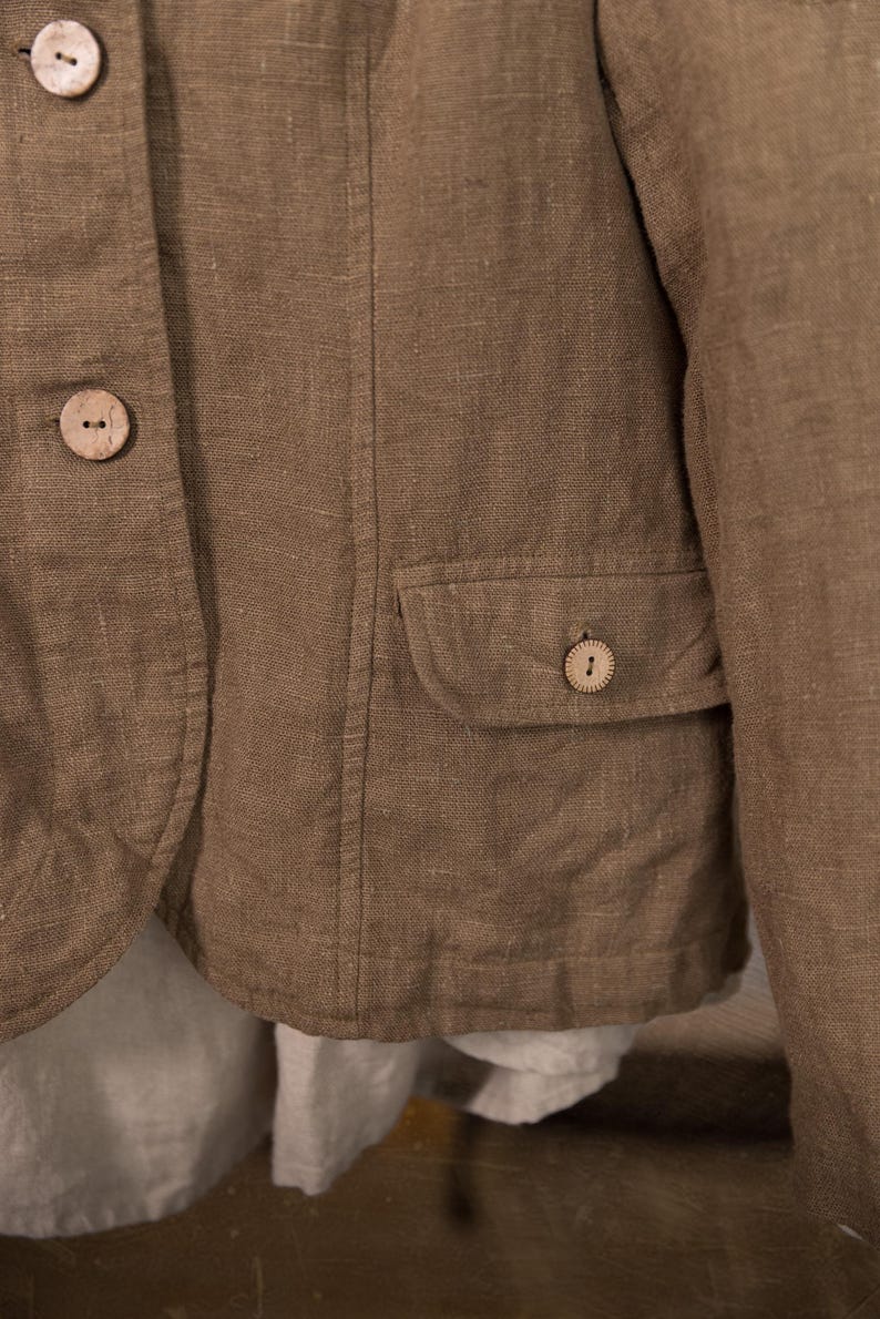A close-up of a brown linen jacket with two wooden buttons and a small pocket. The jacket has a tailored fit and a natural, textured appearance.