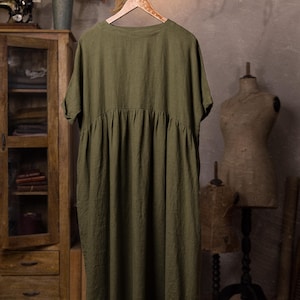 Olive green linen dress shown from the back, hanging from a branch on a decorative wooden hanger, full-length view with round neckline, short sleeves, gathered yoke seam and flowing skirt panel; background includes a wooden cabinet, dried fronds, and mannequins.