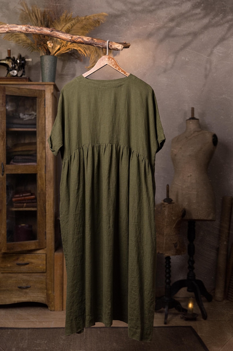 Olive green linen dress shown from the back, hanging from a branch on a decorative wooden hanger, full-length view with round neckline, short sleeves, gathered yoke seam and flowing skirt panel; background includes a wooden cabinet, dried fronds, and mannequins.