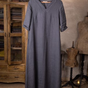 linen dress hanging on hanger from a branch, front view full-length crop, V-slit neckline, short yoke seam lines at chest, elbow-length sleeves with turned cuffs, plain front panel, straight hem; vintage cabinet and mannequin in background, knockknocklinen.