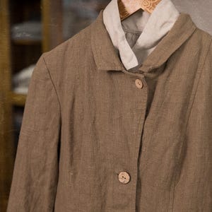 Brown linen jacket on a wooden hanger, shown in a front close-up, with a raised collar, three visible buttons, textured fabric, vertical seams, and a pale linen collar underneath.