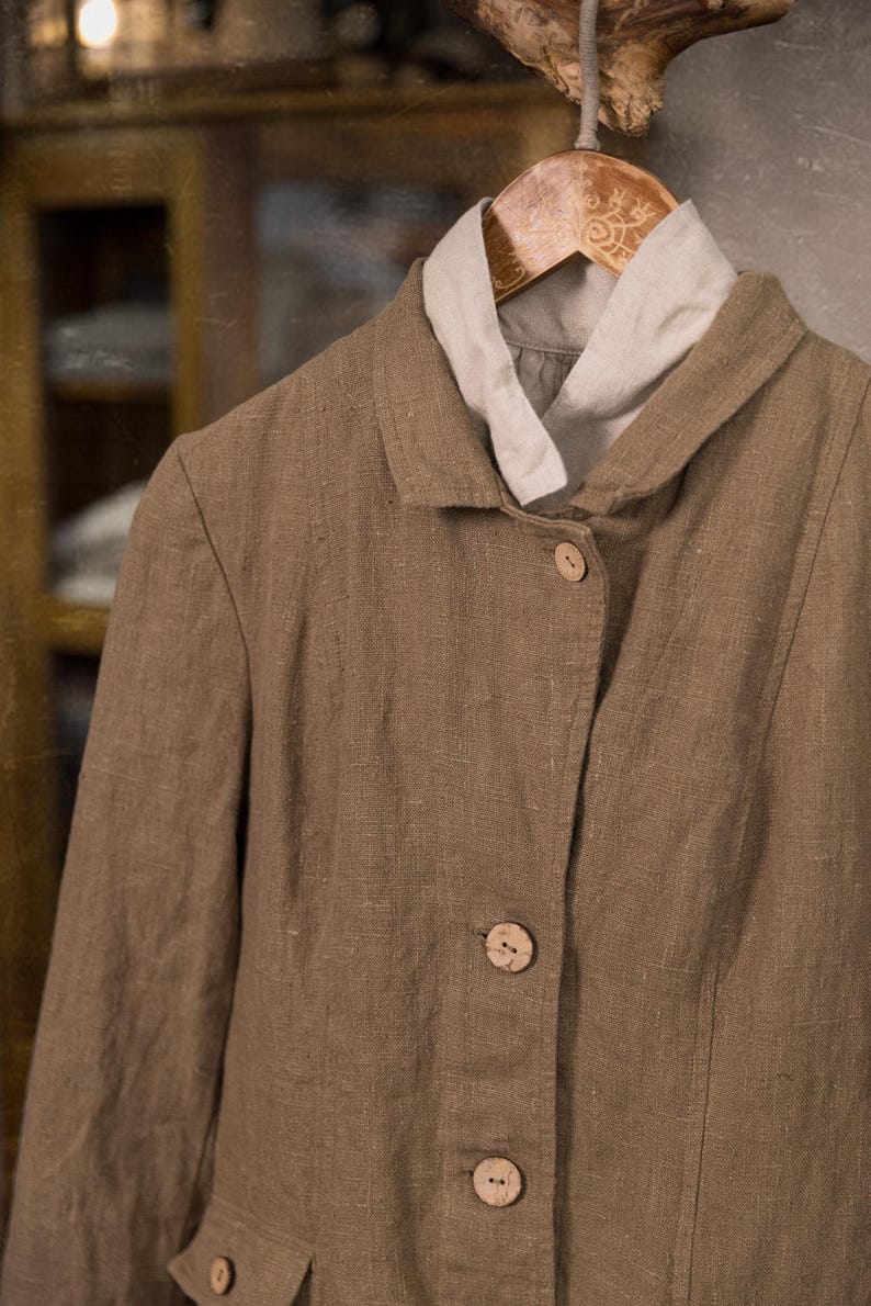 Brown linen jacket on a wooden hanger, shown in a front close-up, with a raised collar, three visible buttons, textured fabric, vertical seams, and a pale linen collar underneath.