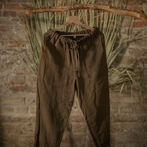 Dark brown-green linen pants with loose fit, gathered elastic waistband, and long center-front drawstring ties, displayed on a hanger in studio.