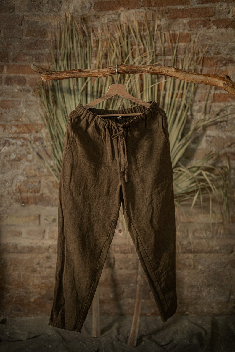 Dark brown-green linen pants with loose fit, gathered elastic waistband, and long center-front drawstring ties, displayed on a hanger in studio.