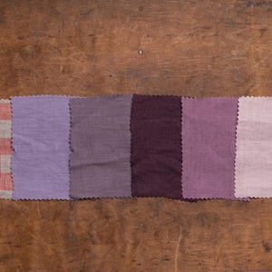 KnockKnockLinen linen fabric samples—6 swatches in purple tones plus red-white gingham; lavender, taupe, deep plum, mauve, pale lilac. Pinked edges, arranged in one row; flat lay on a wooden surface. Fabric swatches / swatch book.