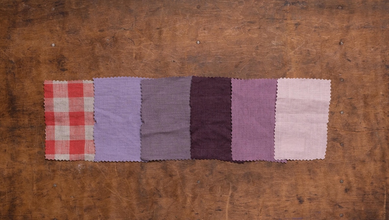KnockKnockLinen linen fabric samples—6 swatches in purple tones plus red-white gingham; lavender, taupe, deep plum, mauve, pale lilac. Pinked edges, arranged in one row; flat lay on a wooden surface. Fabric swatches / swatch book.