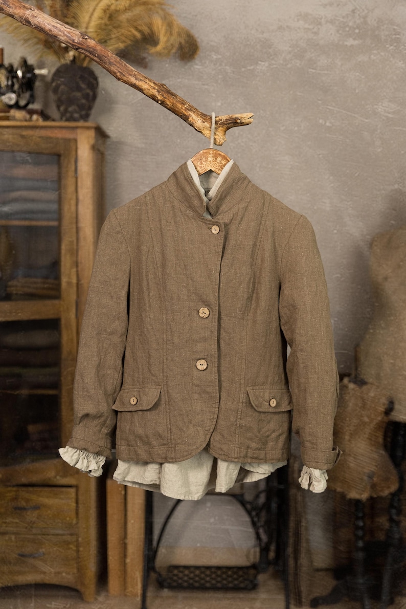 Brown linen jacket on a wooden hanger, shown from the front, with a high collar, three front buttons, flap pockets, long sleeves, and pale linen layers visible at the cuffs and hem.
