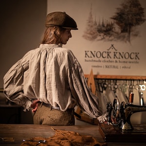 May include: A person wearing a brown cap and a light brown linen shirt stands in front of a sewing machine. The sewing machine is on a wooden table. The wall behind the person has the text "KNOCK KNOCK handmade clothes & home textile timeless | NATURAL | ethical" on it.