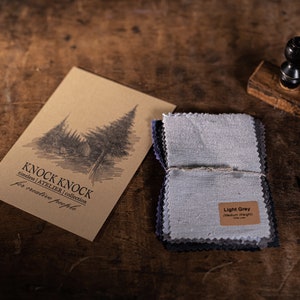 Puede incluir: Un conjunto de muestras de tela en varios tonos de gris, etiquetadas como "Gris claro" y "Peso medio". Las muestras están atadas con un cordel y se encuentran sobre una superficie de madera. También hay una tarjeta de papel marrón con el texto "KNOCK KNOCK timeless | ATELIER | collection for creative people", junto con un sello de madera y unas tijeras.