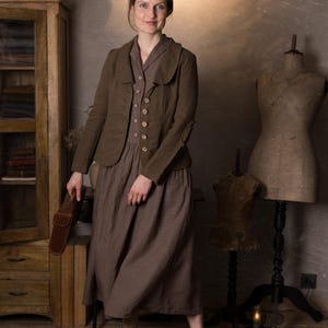Woman in rustic studio wearing mushroom brown slim fit linen jacket with curved collar, long sleeves and button front over brown midi dress, holding brown leather bag, retro linen jacket blazer by knock knock linen.