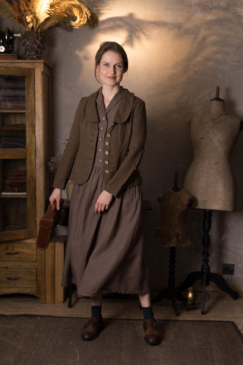 Woman in rustic studio wearing mushroom brown slim fit linen jacket with curved collar, long sleeves and button front over brown midi dress, holding brown leather bag, retro linen jacket blazer by knock knock linen.