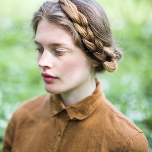 May include: A woman with a braided crown of hair wears a brown linen shirt with a collared neckline. She is standing in a field of green foliage.