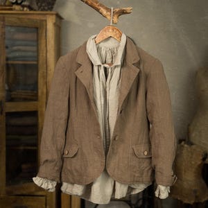 Puede incluir: Una blazer de lino marrón con dos bolsillos delanteros y cierre de botón, combinada con una blusa de color claro. La blazer cuelga de una percha de madera suspendida de una rama natural, lo que sugiere una estética rústica o vintage.