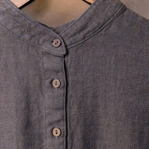 Detail crop of a slate grey linen top hanging on a wooden hanger, showing herringbone weave, mandarin-style collar, top button fastened, and four small buttons down the front placket; neutral backdrop.