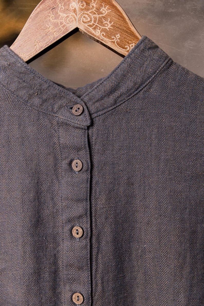Detail crop of a slate grey linen top hanging on a wooden hanger, showing herringbone weave, mandarin-style collar, top button fastened, and four small buttons down the front placket; neutral backdrop.