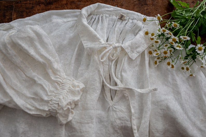 Detail flat lay of a white flax linen smock dress neckline with tie closure and gathered yoke, with a puff sleeve and ruffled cuff on a wooden table.
