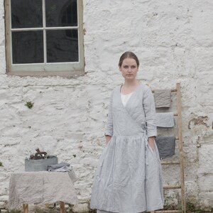 A person wearing a light grey, long-sleeve linen dress with a white undershirt and grey shoes stands in front of a white stone wall with a window. To the left, a small table covered with a grey cloth holds a metal container with various items. To the right, a wooden ladder leans against the wall, draped with folded grey fabrics. The brick-paved ground adds texture to the scene. The text "KNOCK KNOCK LINEN" is visible at the bottom left.