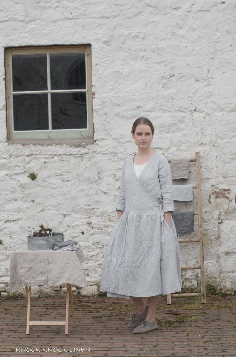 A person wearing a light grey, long-sleeve linen dress with a white undershirt and grey shoes stands in front of a white stone wall with a window. To the left, a small table covered with a grey cloth holds a metal container with various items. To the right, a wooden ladder leans against the wall, draped with folded grey fabrics. The brick-paved ground adds texture to the scene. The text "KNOCK KNOCK LINEN" is visible at the bottom left.