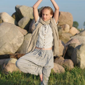 Person standing in tree pose wearing a dusty sky buttoned linen top with short sleeves and loose linen pants. Arms raised overhead with palms together. Photographed outdoors on rocks with greenery. Buttoned blouse, linen summer shirt, loose linen top.