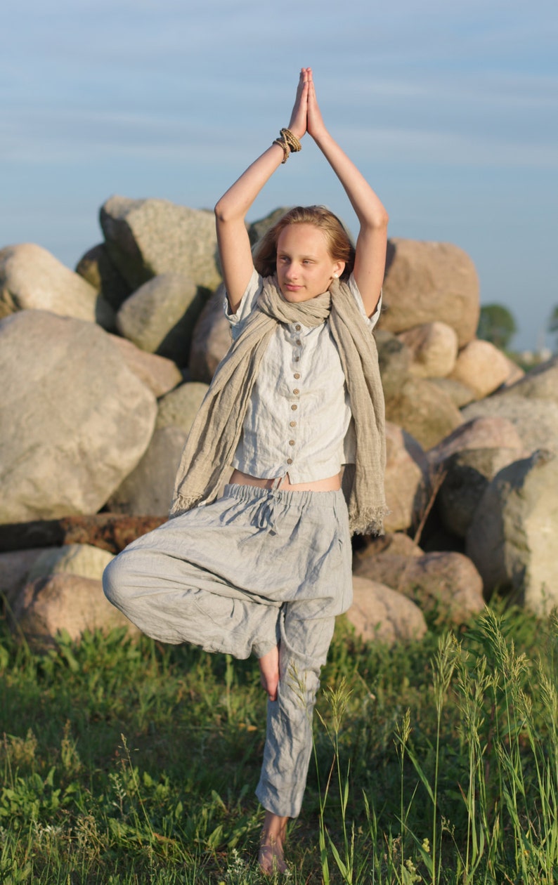Person standing in tree pose wearing a dusty sky buttoned linen top with short sleeves and loose linen pants. Arms raised overhead with palms together. Photographed outdoors on rocks with greenery. Buttoned blouse, linen summer shirt, loose linen top.