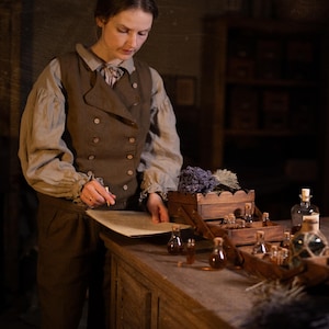 Brown linen waistcoat on model, waist-up view; sleeveless with two rows of light buttons and curved overlap; worn over pale blouse with gathered sleeves and ruffled cuffs; pencil and paper on counter with corked vials and lavender.