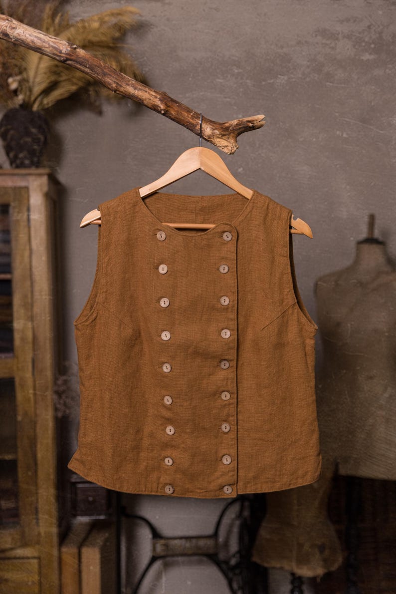 SPARROW linen vest in brown, displayed on a hanger in a front-view full-length photo, with a round neckline, sleeveless armholes, double rows of light buttons, an offset front panel, and a slightly curved hem, Knock Knock Linen.