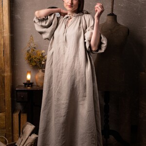 Natural flax linen dress worn by a model, full-length front view, tie strings at gathered neckline, puffed sleeves with ruffled cuffs, loose straight silhouette, wrinkles visible across front, ankle-length hem; mannequin torso on right.
