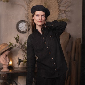 Waist-up to mid-thigh view of a model wearing a black linen shirt with stand collar, long sleeves, light buttons, and a chest pocket, black beret visible, sewing table and wall clock in background, KnockKnockLinen studio