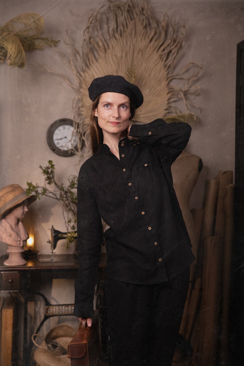 Waist-up to mid-thigh view of a model wearing a black linen shirt with stand collar, long sleeves, light buttons, and a chest pocket, black beret visible, sewing table and wall clock in background, KnockKnockLinen studio
