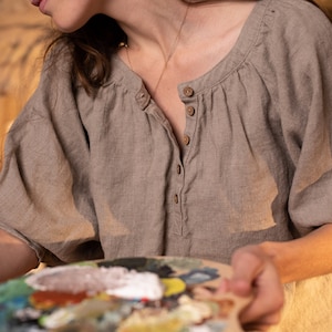 Person wearing one loose-fit beige linen shirt with puff sleeves and front buttons, holding painter’s palette with multiple paint colors. Arm extended in painting gesture. Rustic indoor setting with wood textures. Handmade linen clothing, artistic context.