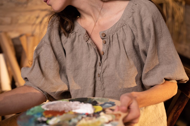 Person wearing one loose-fit beige linen shirt with puff sleeves and front buttons, holding painter’s palette with multiple paint colors. Arm extended in painting gesture. Rustic indoor setting with wood textures. Handmade linen clothing, artistic context.