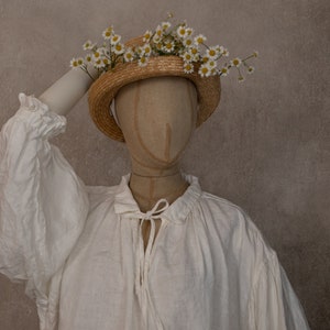 White linen dress displayed on a mannequin, showing the long sleeve and tie neckline, photographed against a plain wall with a straw flower hat.