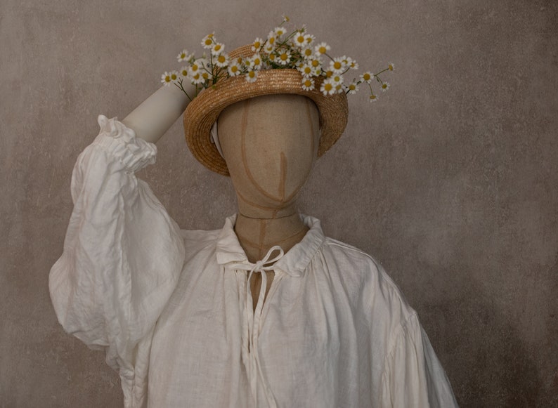 White linen dress displayed on a mannequin, showing the long sleeve and tie neckline, photographed against a plain wall with a straw flower hat.